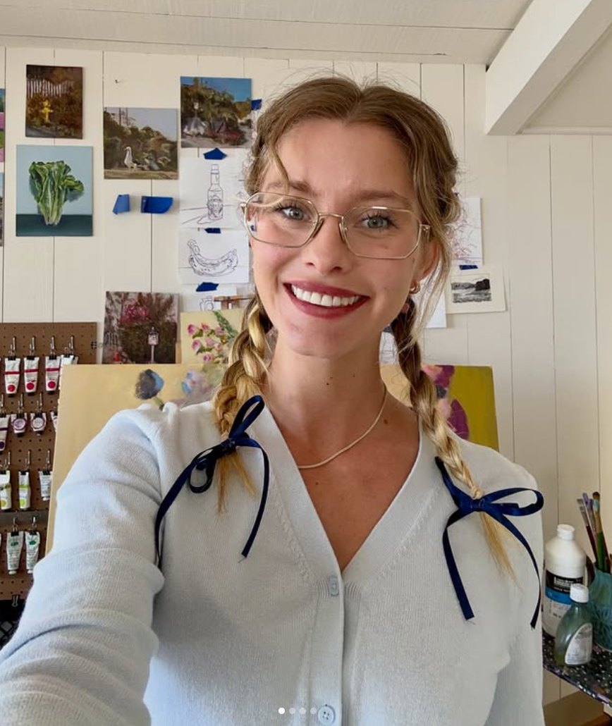 Haven smiling in glasses and braids with navy bows, in her painting studio