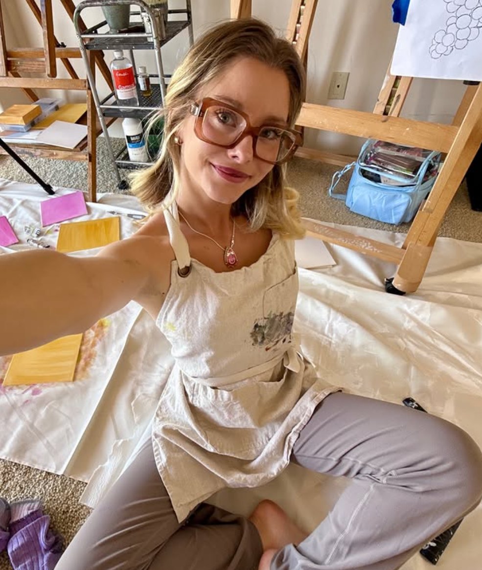 Haven in a paint-splattered apron and glasses, sitting among canvases and easels